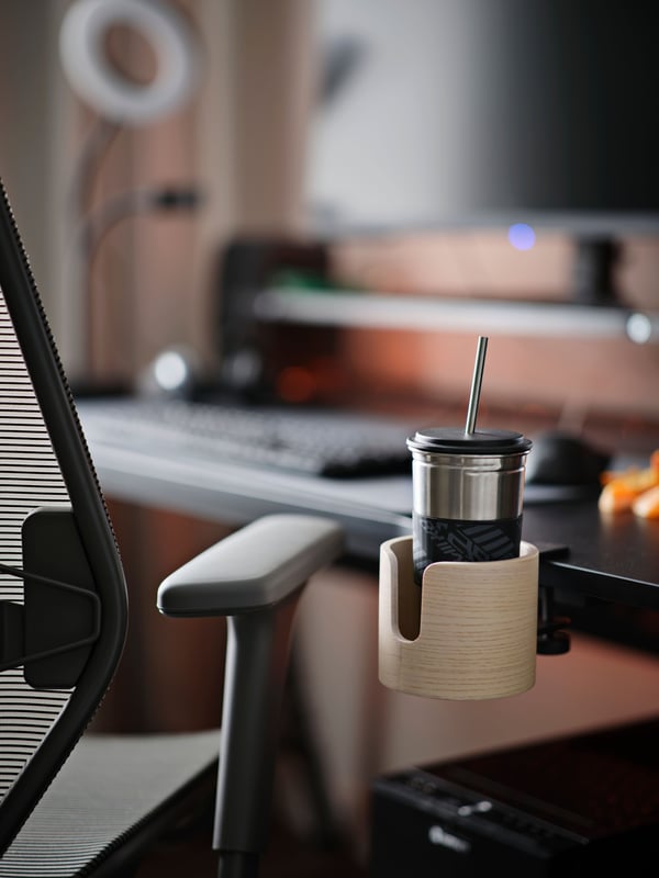 An ash veneer LÅNESPELARE mug holder is clamped onto the edge of a black gaming desk, holding a stainless steel mug and straw.