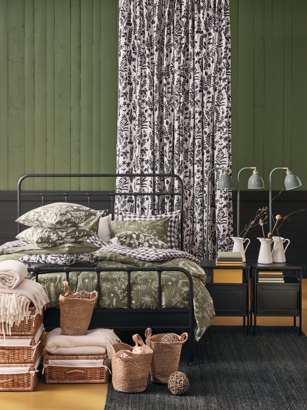 An anthracite STJÄRNÖ bed dressed with SLÅNGULDMAL and EKTANDVINGE duvet covers. Rattan boxes and jute bags stands in front.