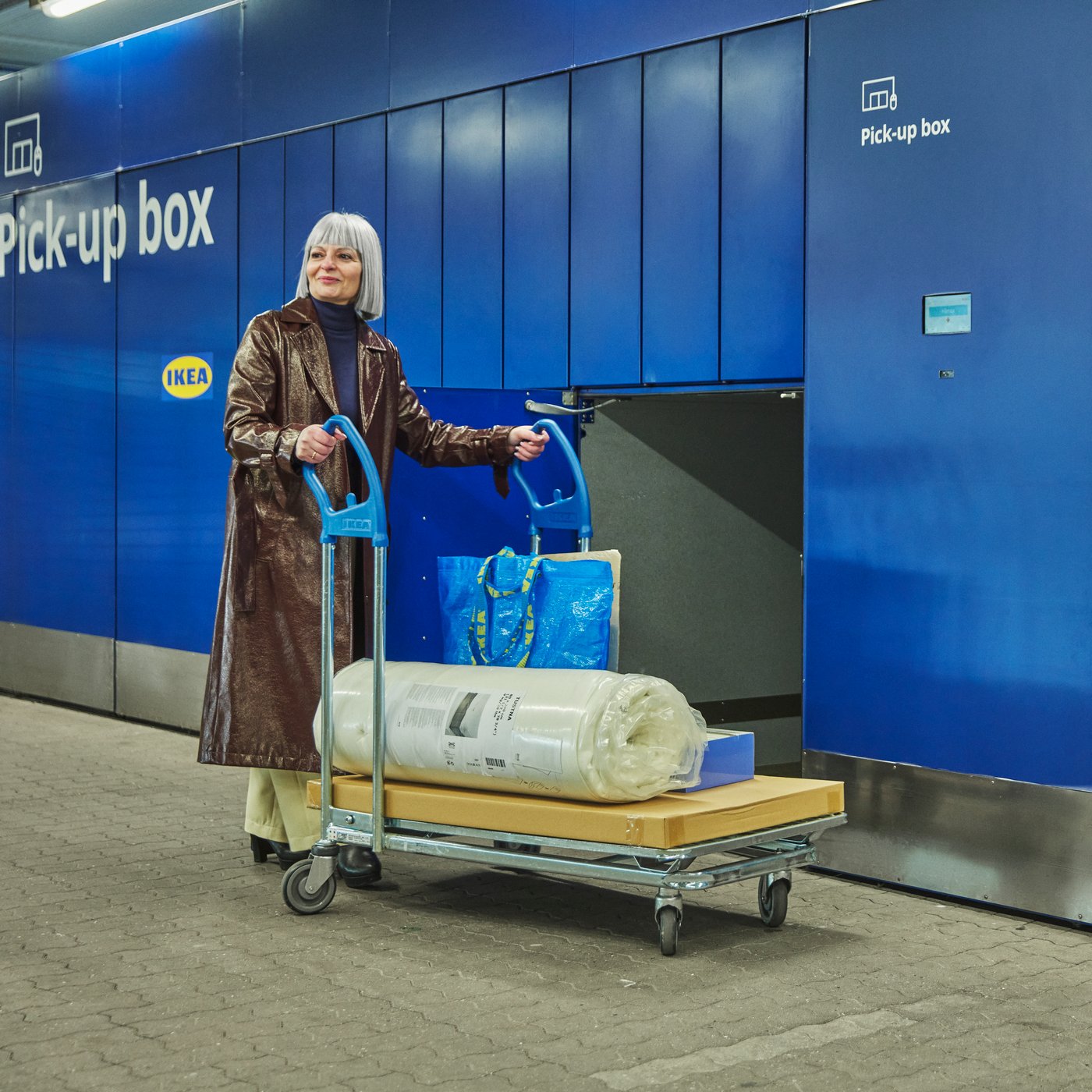 Eine Kundin vor einer Click & Collect Abholbox, wo sie gerade ihre Bestellung auf einem Flachwagen abgeholt hat. 