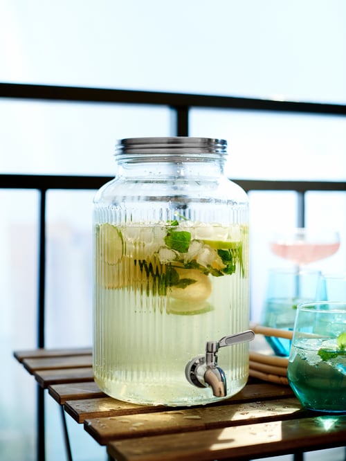 Ce bocal en verre VARDAGEN de 5 litres avec couvercle et robinet contient une boisson aux agrumes. Il est prêt à verser des boissons sur une table de balcon.
