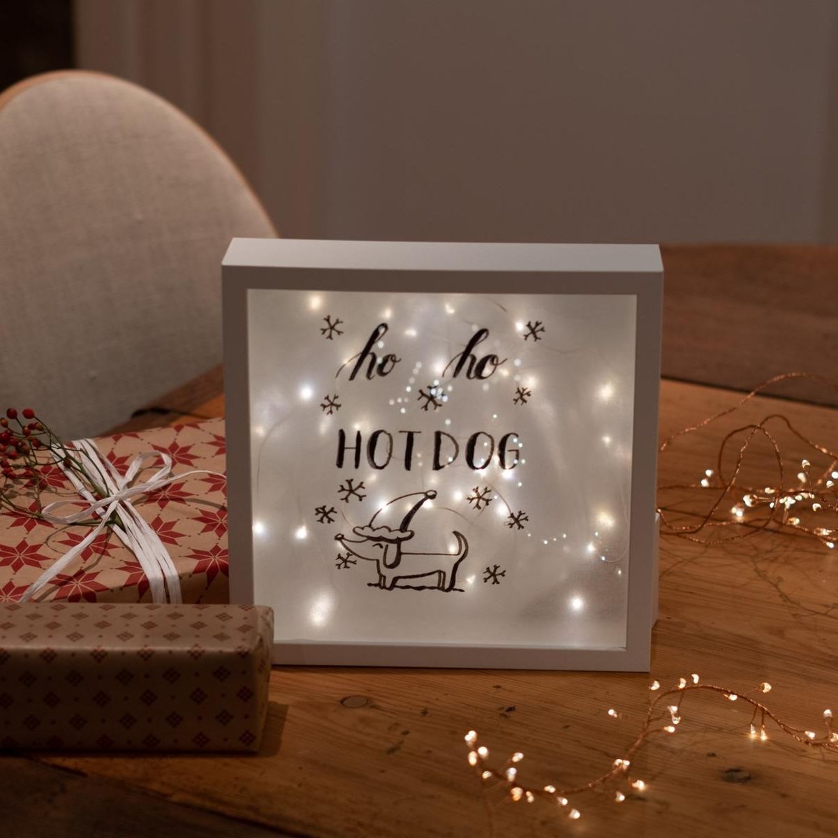 Leuchtbox mit Schriftzug ‚ho ho HOT DOG‘ und Hundemotiv auf Holztisch, daneben Geschenke und Lichterkette.