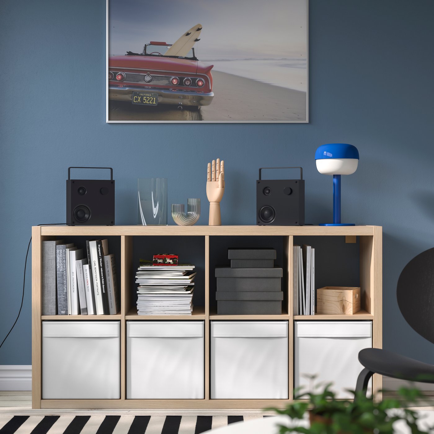 Modulo contenitore KALLAX in legno chiaro con vani aperti pieni di riviste e scatole e tre contenitori bianchi in basso, contro una parete blu con quadro e lampada da tavolo.