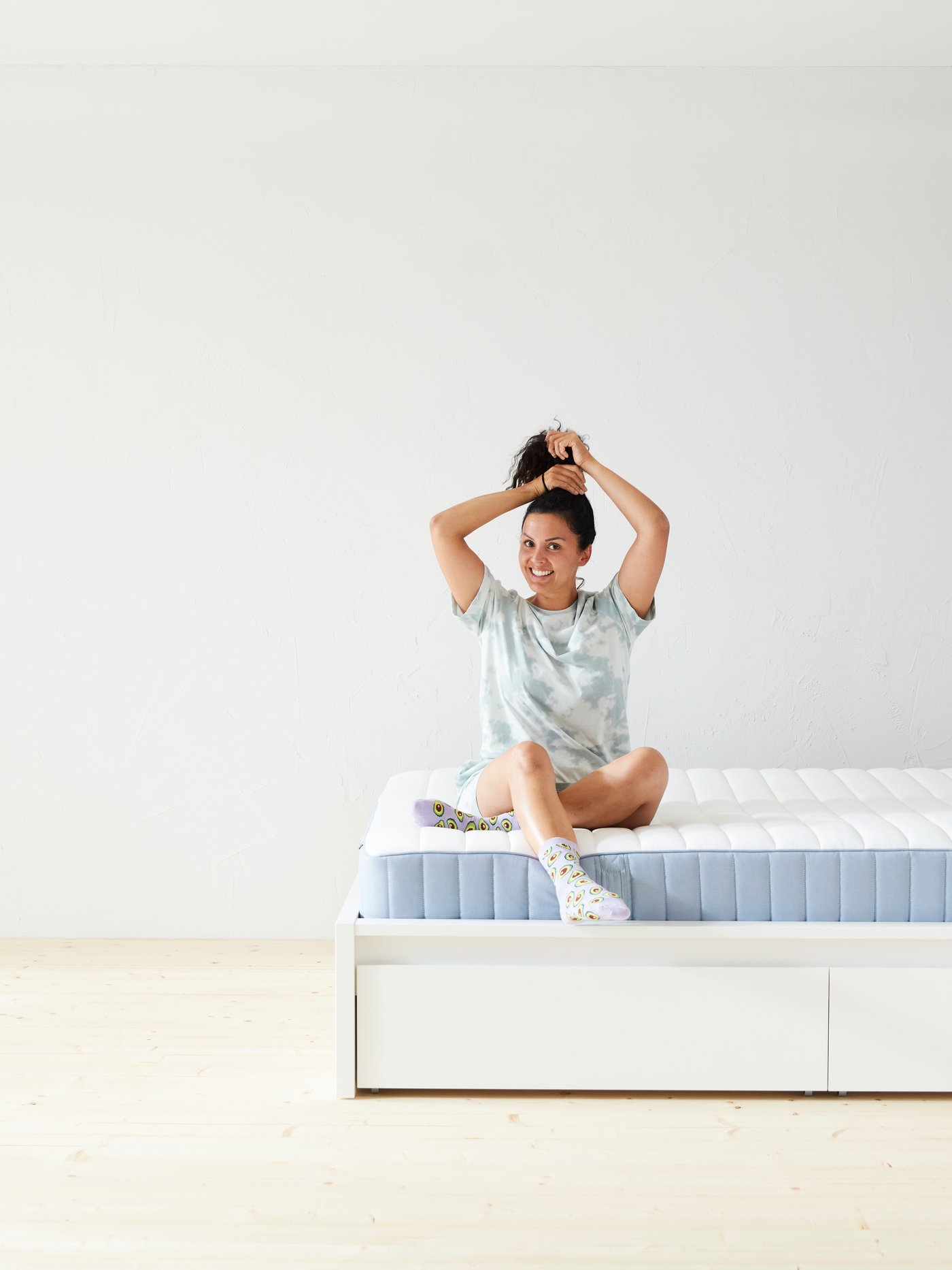 A woman sits on a bare mattress, looks happily at the camera and fixes her hair