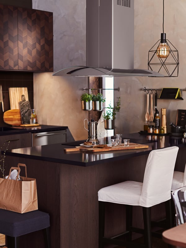 A dark wood kitchen with an island that has a BRUNSTA pendant lamp overhead. A coffee pot and glasses are on the counter.