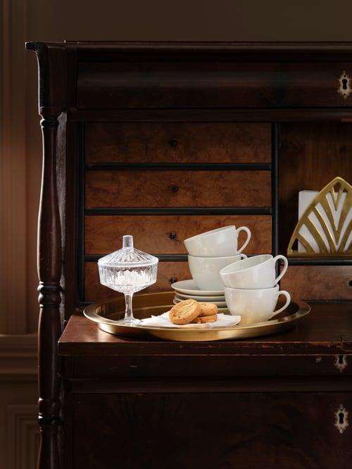 Des tasses et des soucoupes VÄRDERA blanches, empilées sur un plateau en or avec un sucrier en verre taillé, des biscuits et un porte-serviette en or.