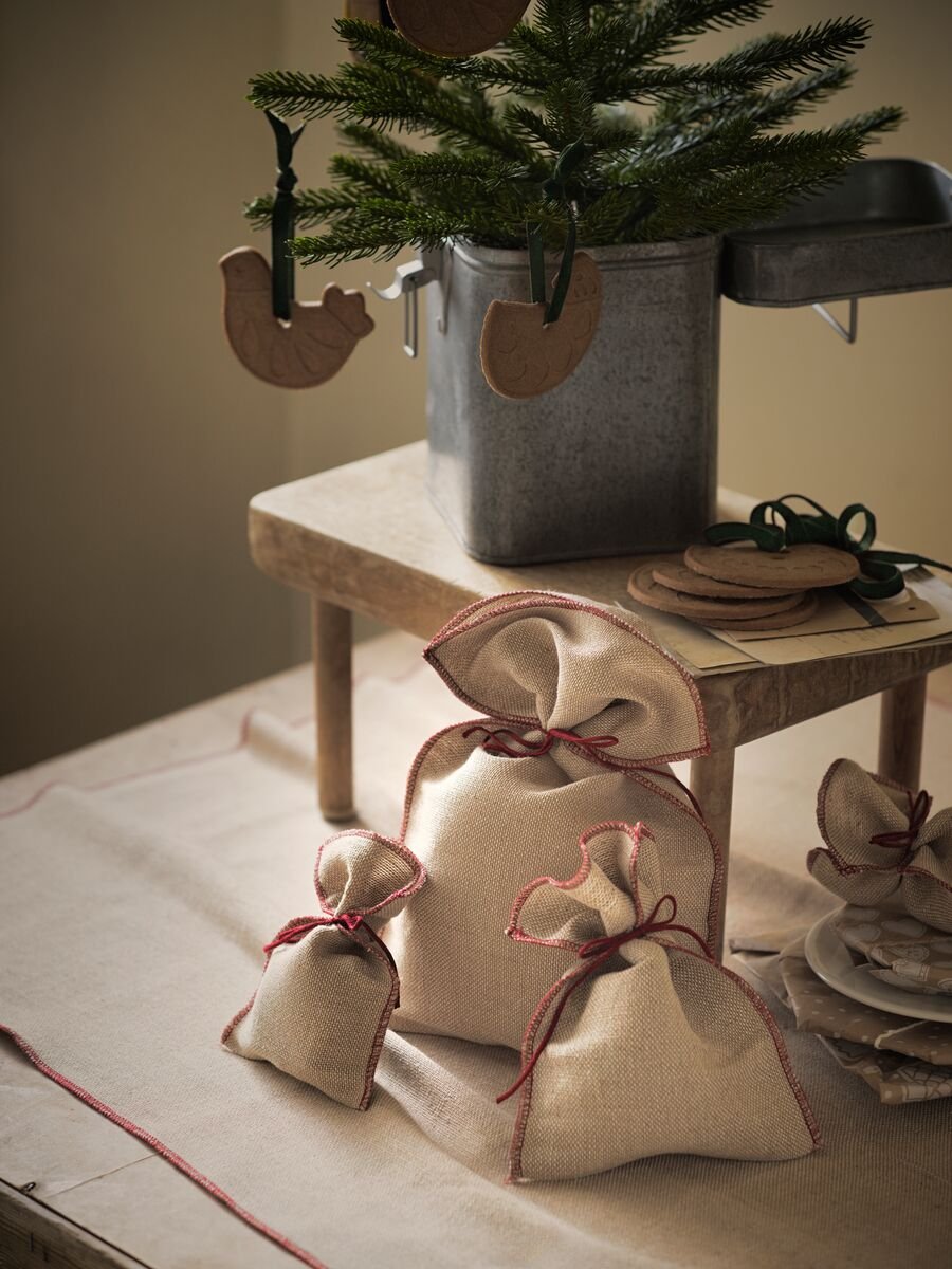 A number of gifts wrapped in VINTER 2020 jute gift wrap underneath a small pine tree with wooden hanging animals on it.