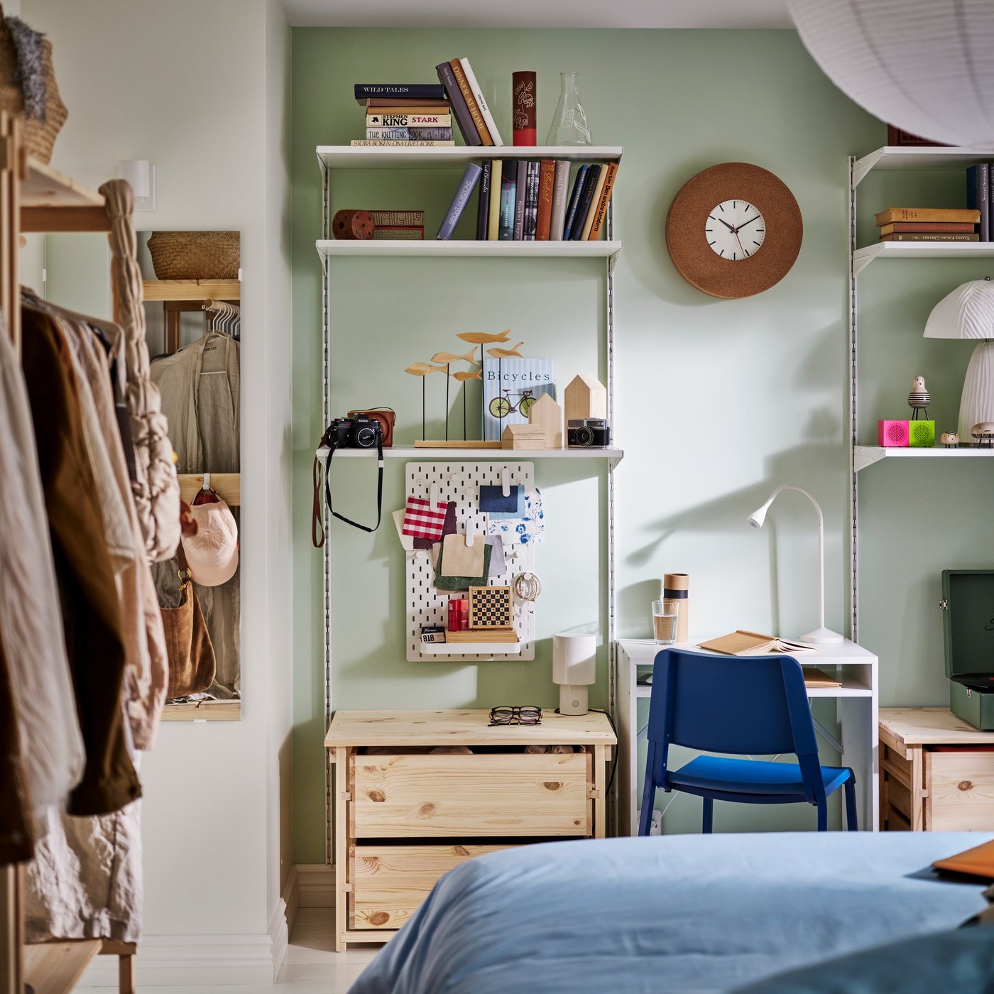 Ett BOAXEL förvaringssystem med hyllor och pegboard uppsatt mot en ljusgrön vägg bredvid ett litet skrivbord och en blå stol.