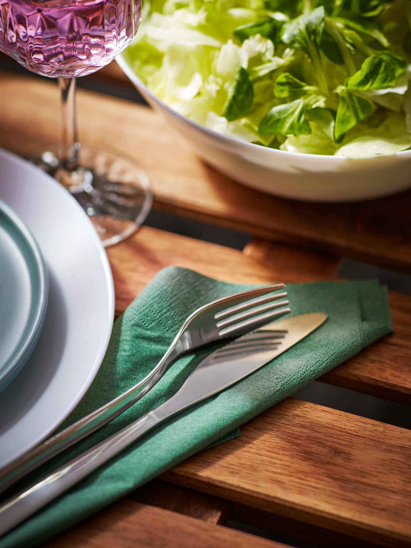 Cubiertos en una servilleta de papel verde, junto a una ensaladera con ensaladas, una copa de vino y un plato blanco.