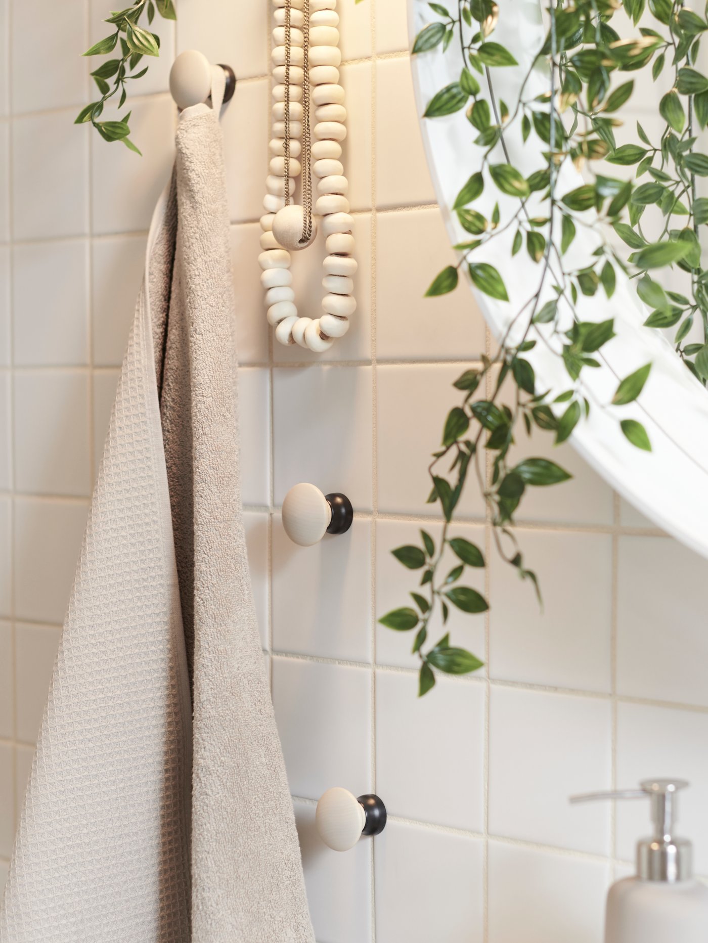 A beige SALVIKEN bath towel and necklaces hanging beside a mirror on LILLASJÖN birch knobs in a bathroom with white tiles.