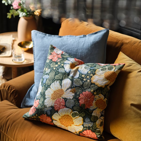 A couple of cushions lean on a yellow-brown sofa, one is dressed in an ARENBOK cushion cover with a floral pattern.