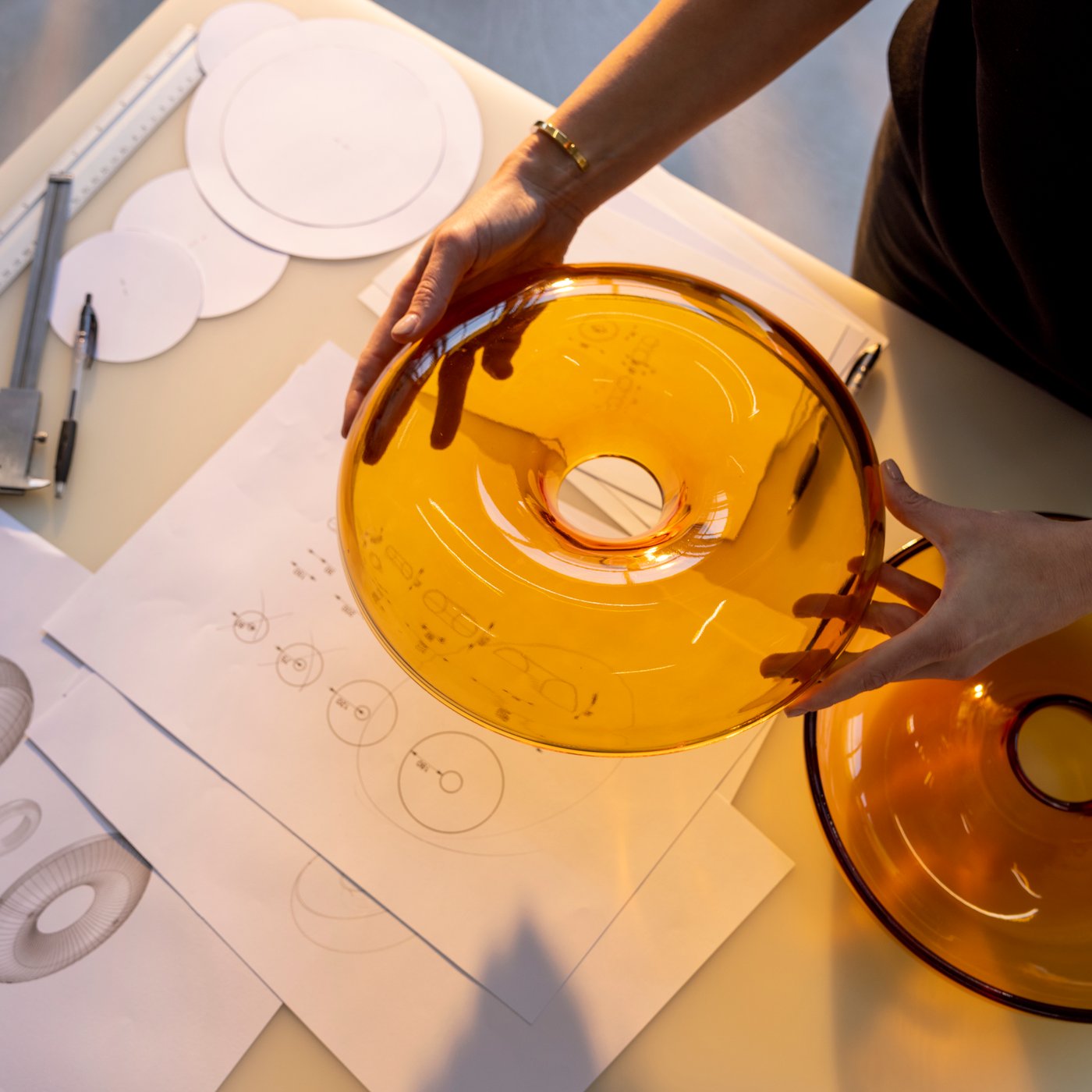 A pair of hands holds up a lid of an orange VARMBLIXT glass doughnut-shaped bowl above a table with sketches of the object.