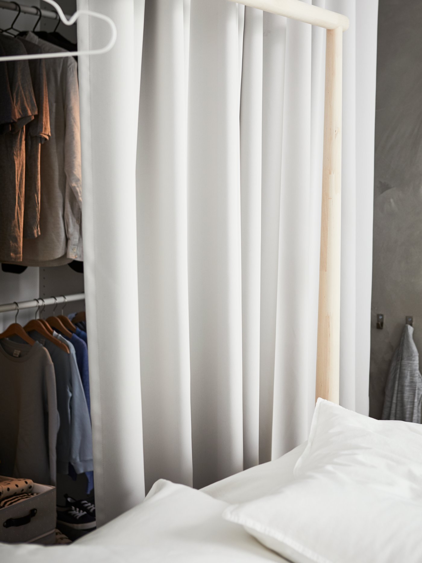 Light grey MAJGULL room darkening curtains, used as a divider between a closet full of clothes and a birch GJÖRA bed frame.