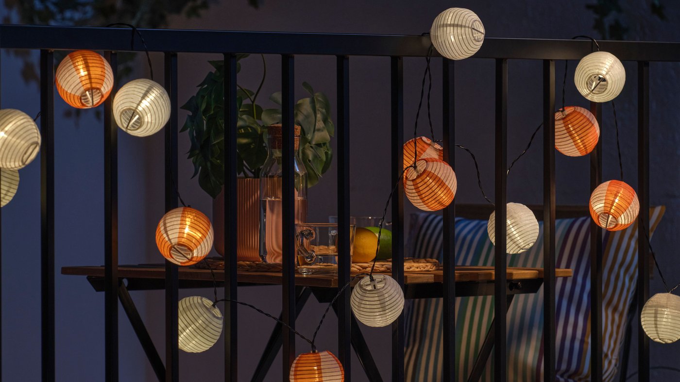 Catena luminosa da esterno SOLVIDEN con piccole lanterne arancioni e bianche appese alla ringhiera di un balcone, che illuminano un tavolino con cuscini a righe e bevande per creare un angolo accogliente nelle sere d’estate.