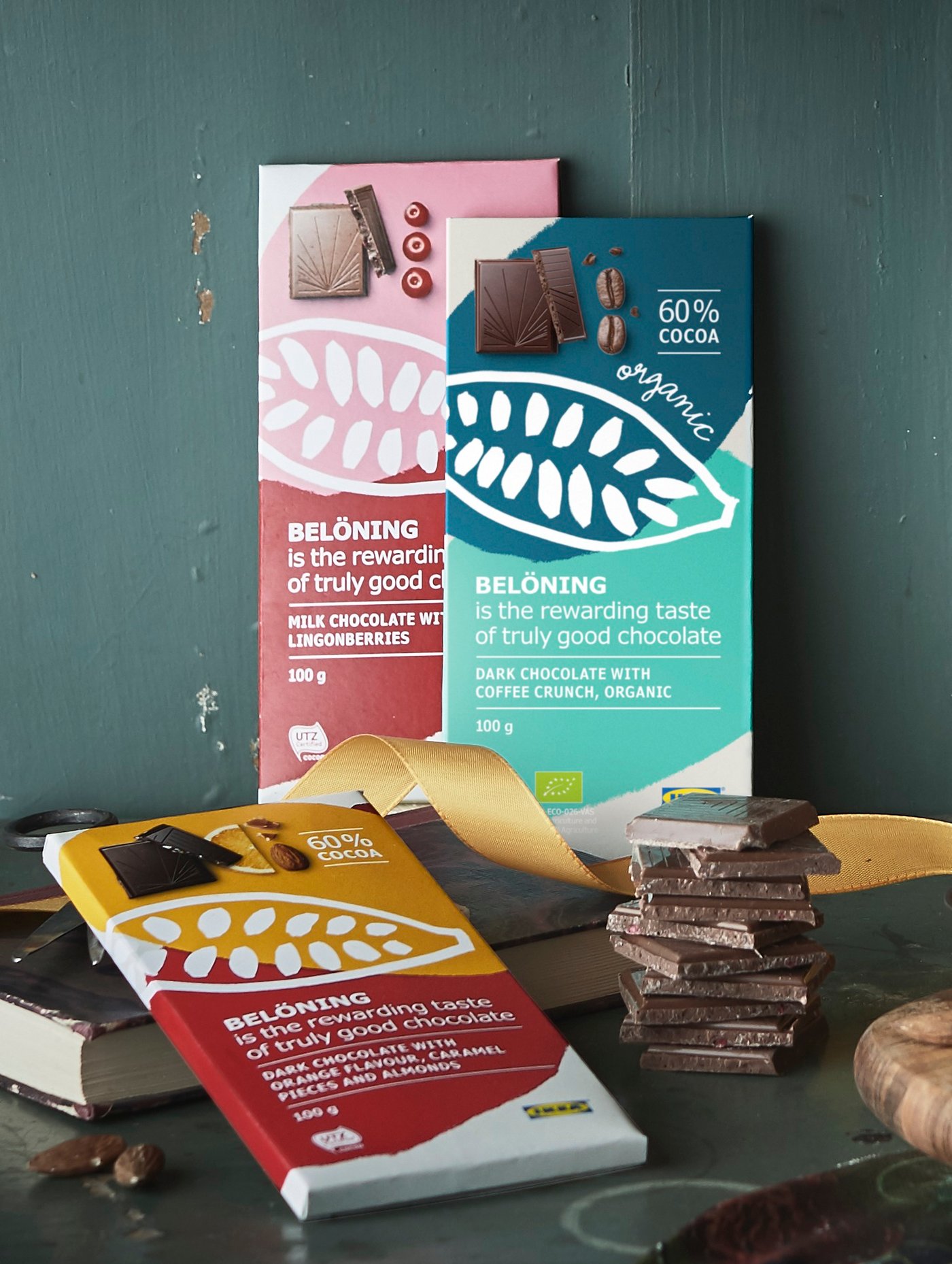 Two BELÖNING chocolate bars leaning against a green wall, another bar is laying on top of a book and a pair of scissors.