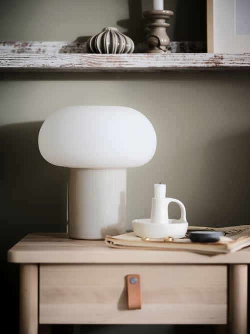 Une lampe de table DEJSA blanc/opalin se trouve à côté d’un bougeoir blanc, sur une table de chevet BJÖRKSNÄS.