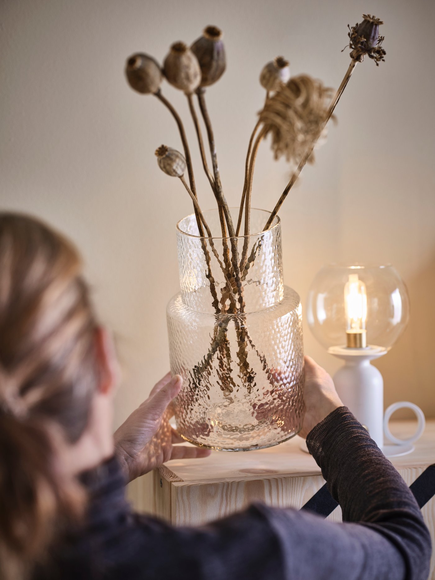 Kobieta ustawiająca wazon KONSTFULL z suszonymi rośłinami na drewnianej szafce.