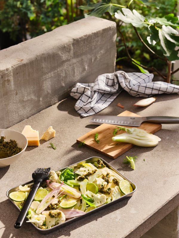 An outdoor concrete worktop with cooking accessories, including a GRILLTIDER barbecue tray filled with sliced vegetables.