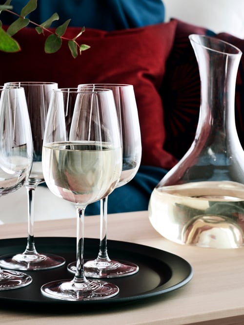 Dining area with white wine glasses, carafe, serving plate, on wooden table, with cushions, plant and sofa in the background.