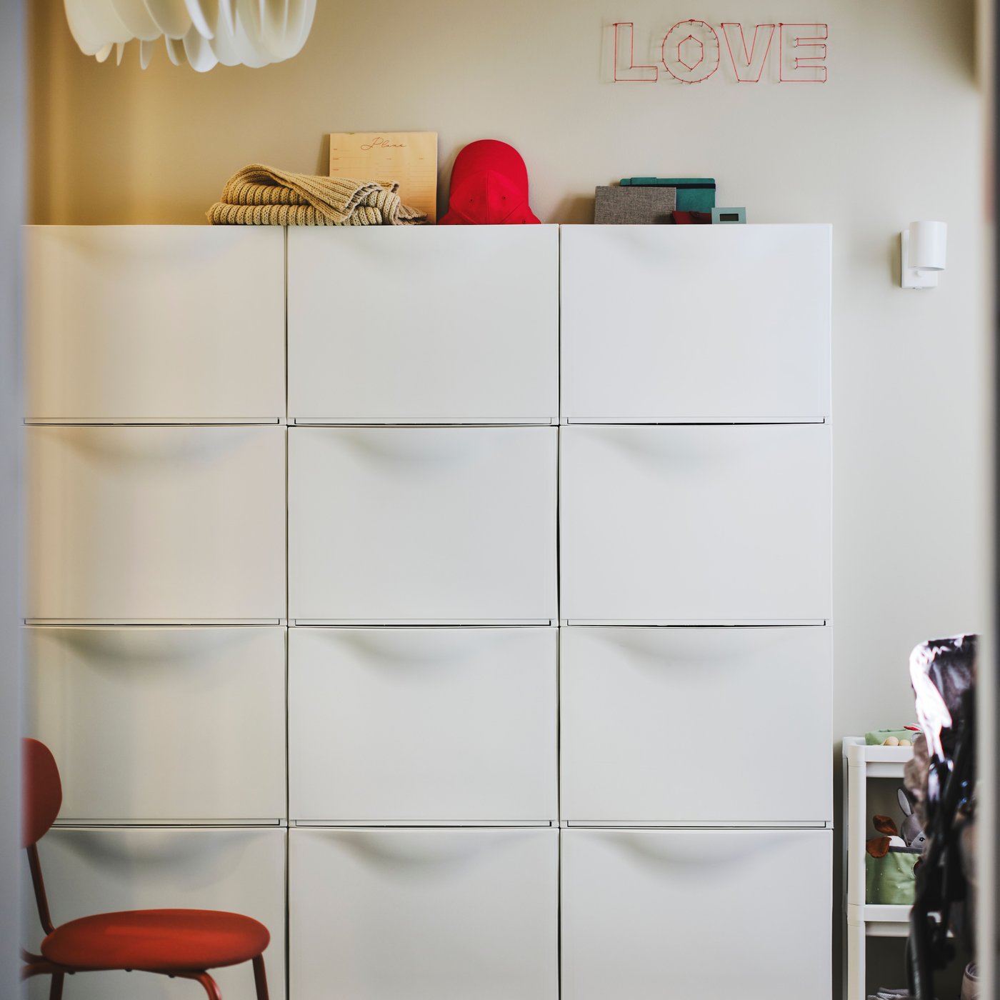 Sex vita TRONES skoskåp på en vägg, med en rödbrun ÖSTANÖ stol framför och en röd hatt och en beige halsduk ovanpå.