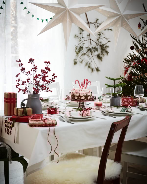 Festively set table with white tablecloth and red VINTERFINT decorative elements. From the ceiling hangs a white STRÅLA lampshade. In the foreground is a brown PINNTORP chair with a fur for a soft seat.