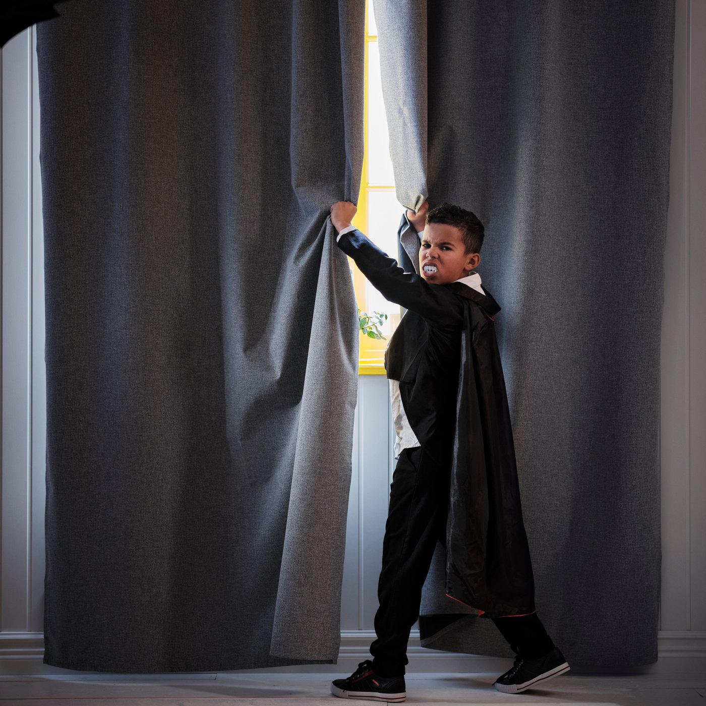 A boy in a Dracula costume stands by a window with black ROSENMANDEL blackout curtains.