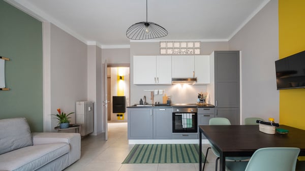 Una cucina/soggiorno con cucina lineare arredata con mobili ENHET, la lavastoviglie e il piano a induzione. A destra ci sono un tavolo SANDSBERG, abbinato alle sedie impilabili LIDÅS con sedile sagomato e schienale flessibile. A sinistra, un divano KIVIK e un tappeto STOENSE blu.