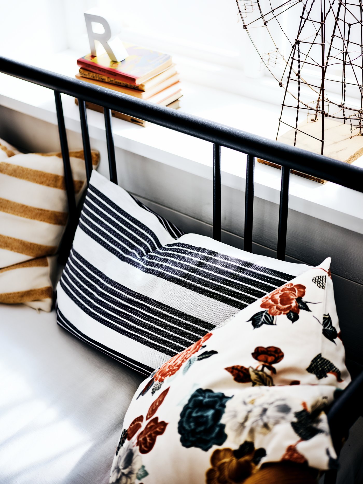 The black metal frame of a day-bed by a window sill with cushions on it that have striped and flower-patterned covers.