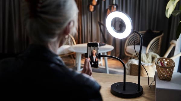 A woman sits at a desk, in the process of making a video recording using a LÅNESPELARE ring light with phone holder.