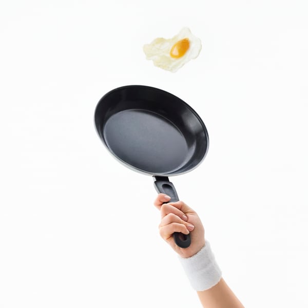 An arm is holding a TAGGHAJ frying pan as if it were a tennis racquet, catching a fried egg in the air as if it were a ball.