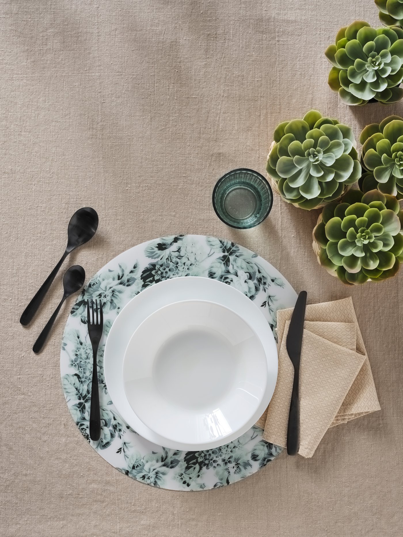Vista aérea de un plato hondo OFTAST blanco sobre un plato más grande y un mantel individual OFTAST. También se muestran cubiertos, servilletas, un vaso y plantas.