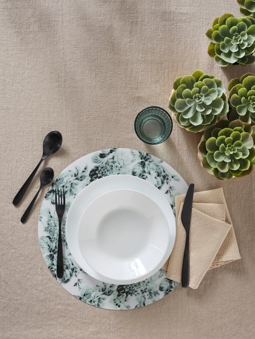 Une vue aérienne d’une assiette OFTAST blanche placée sur une assiette plus grande et un napperon. Près de l’assiette se trouvent des couverts, des serviettes de table, un verre et des plantes.