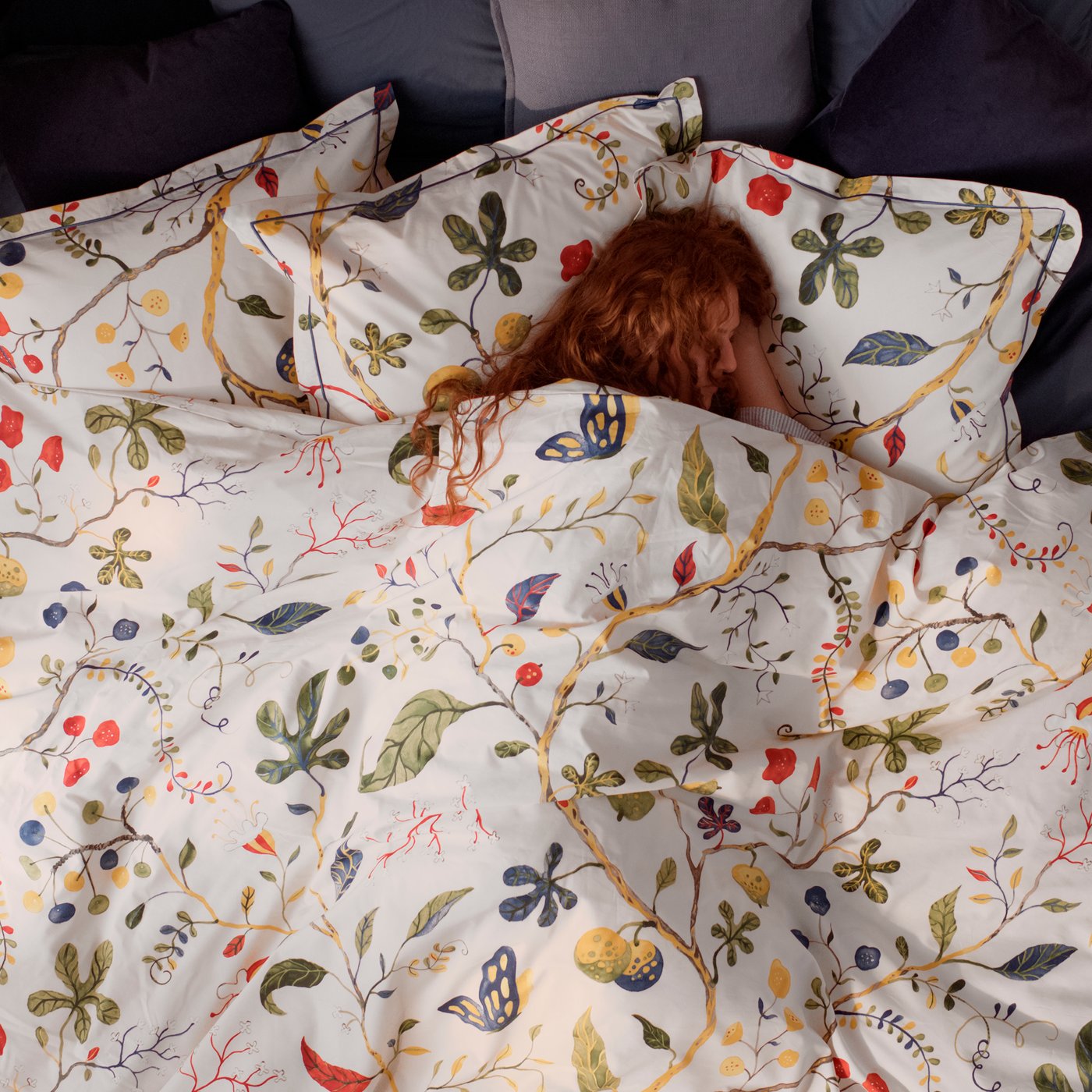 A person with red hair is sleeping in a bed underneath the light beige KRONBUSKE bed linen with a floral pattern.