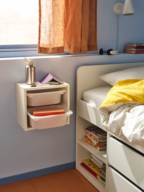 A white TROFAST wall storage with two white storage boxes in it beside a bed. A water bottle and a book are on top.