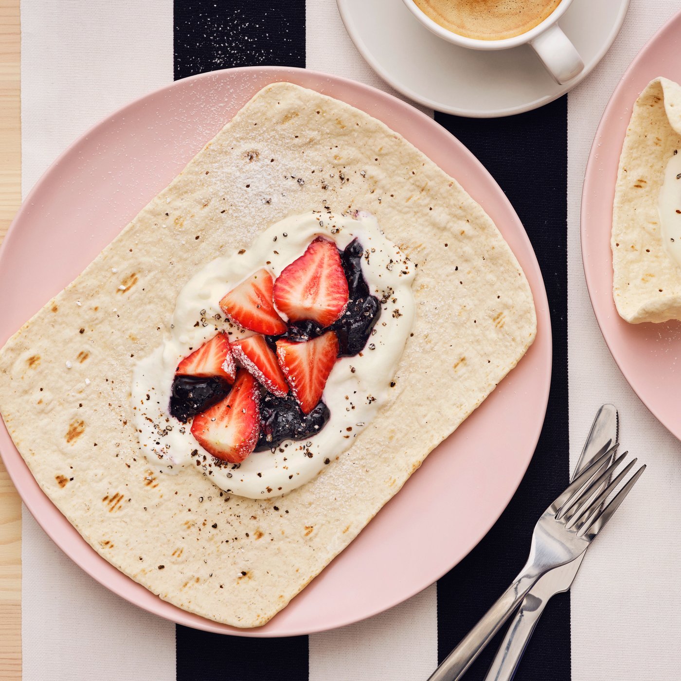 A pink plate, with a piece of flatbred with berries and whipped cream, is placed on a black and white striped surface.
