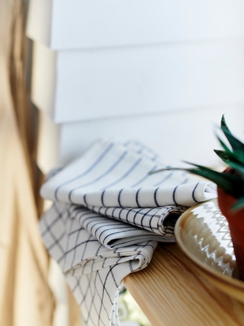 A pile of folded RINNIG tea towels with different patterns sit at the edge of a wooden surface in a kitchen.