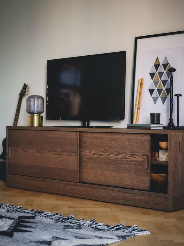 A brown-stained oak veneer TONSTAD TV bench with a TV and framed artwork on top and a grey and black patterned rug in front.