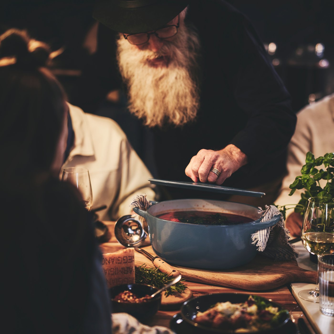 Un homme âgé avec une longue barbe soulevant le couvercle d’une cocotte, pendant que les convives assis à table le regardent.