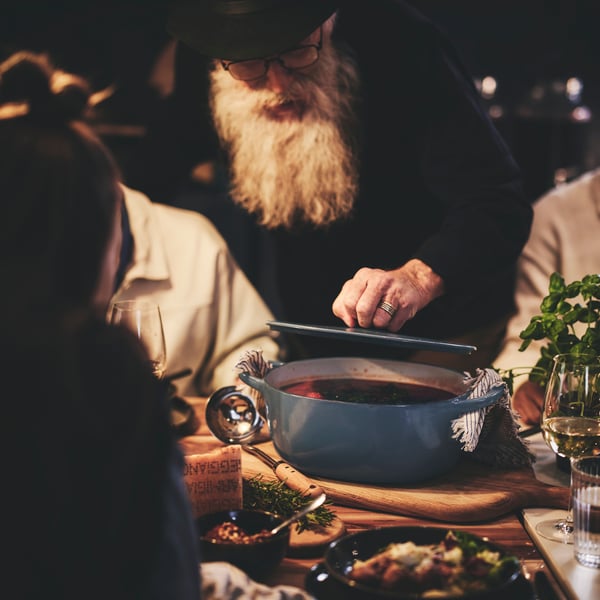 An elderly man with a long beard lifts the lid on a casserole dish while dinner guests seated at the table look on.