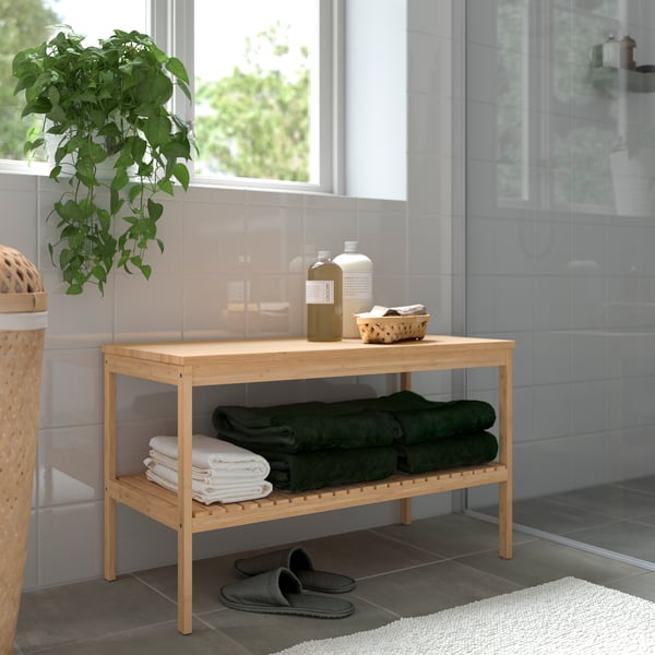 A bamboo RÅGRUND bench storing towels and shampoo is under a window by a shower. Slippers and a rug are near the bench.