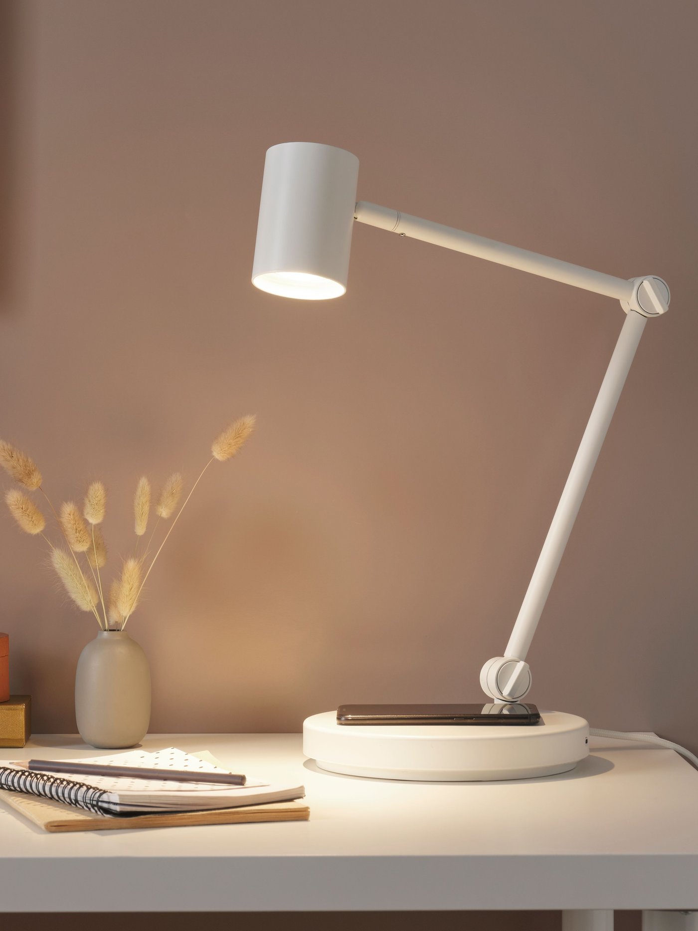 스타일리시한 책상 코너가 있고 거기에 놓인 화이트 NYMÅNE 뉘모네 독서등의 하단에 있는 무선충전기에 스마트폰이 놓여 있는 모습.