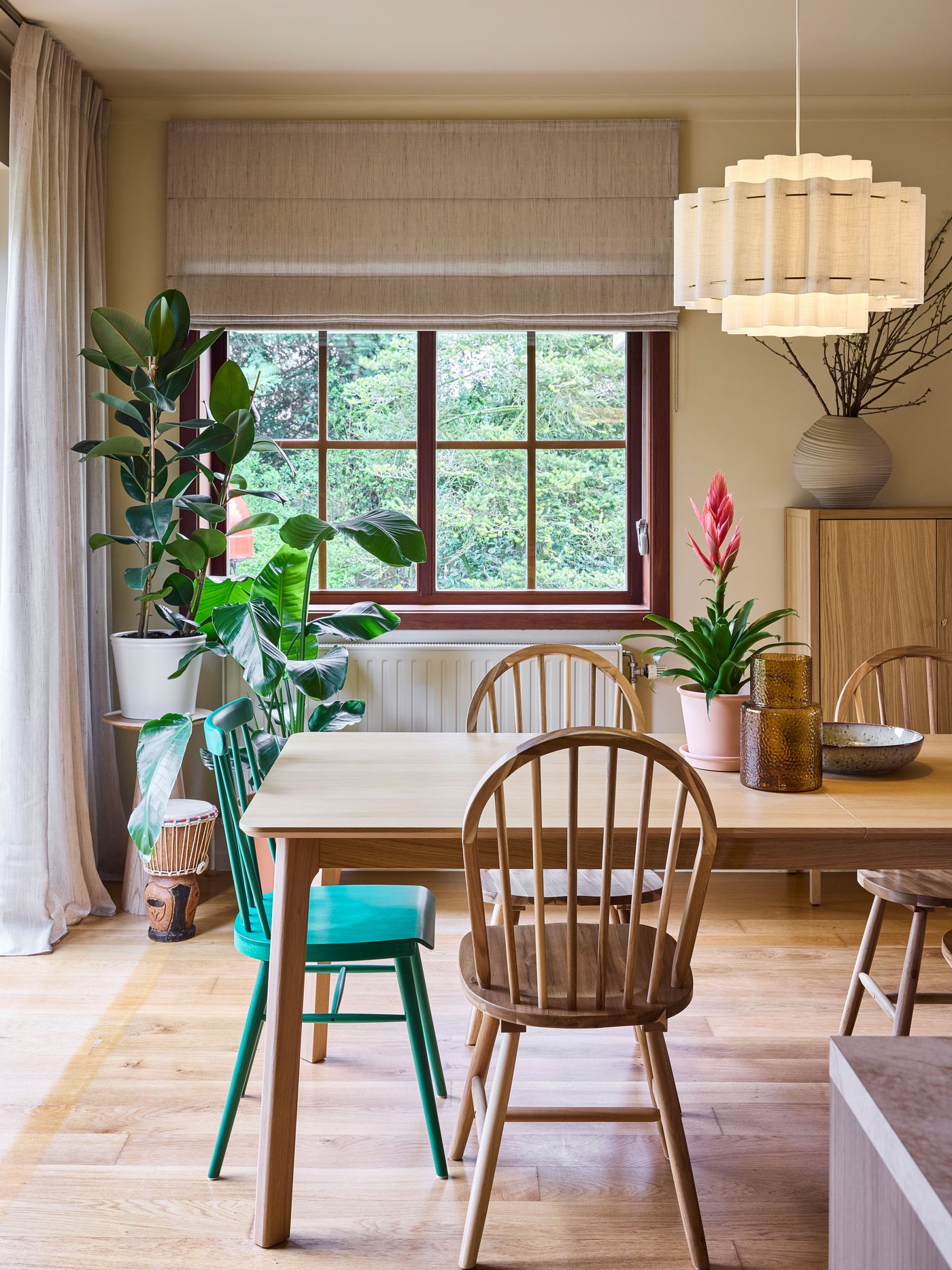 Eetruimte met houten tafel, mix van stoelen, groene plant bij raam, beige rolgordijn en hanglamp voor een warme, huiselijke sfeer.
