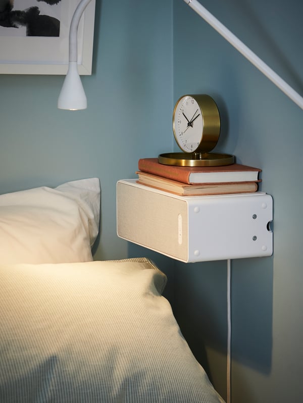 White, SYMFONISK WiFi speaker mounted horizontally on a blue wall by a bed, with two books and a gold-coloured clock on top.