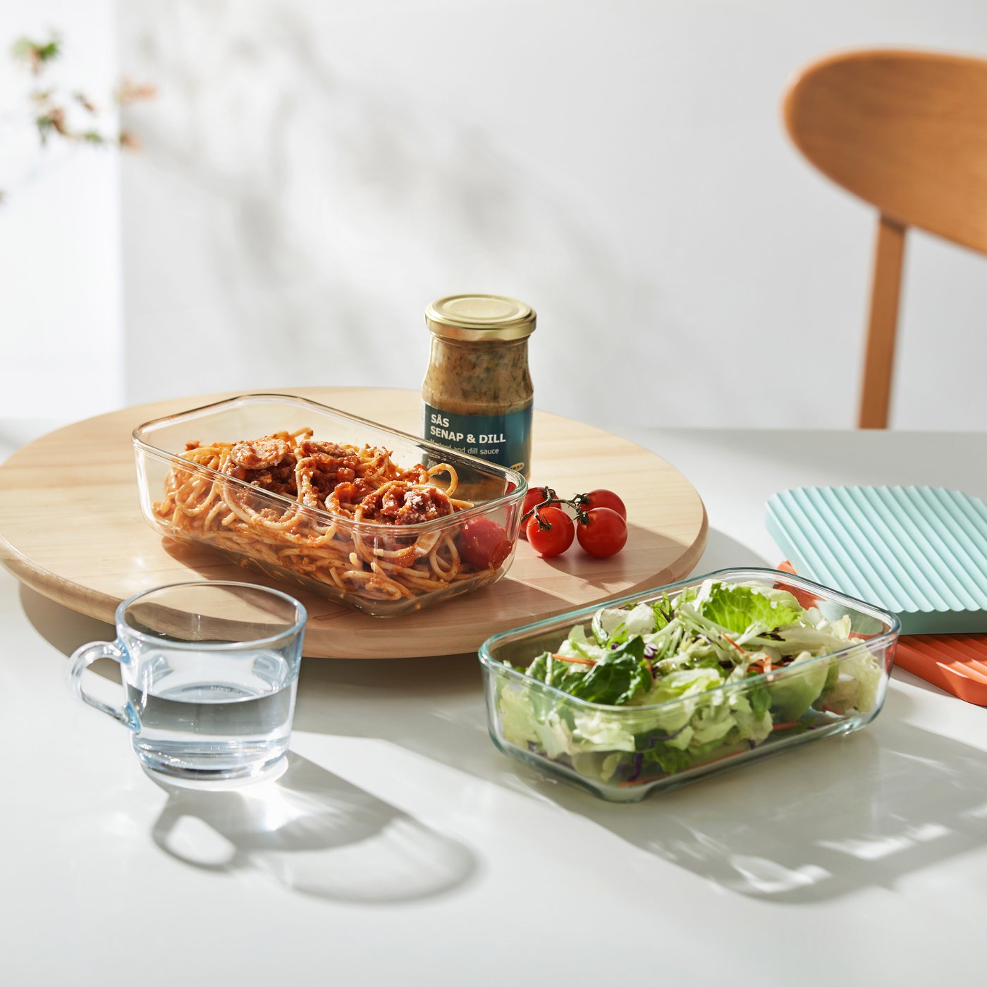 Clear glass storage containers filled with food sitting on a dining table with a mug of water. 