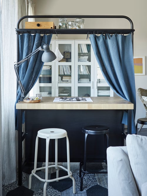 Living room with kitchen island with rack, bar stools, work lamp, fabric, rug, glasses, storage unit, sofa, curtains, frames.