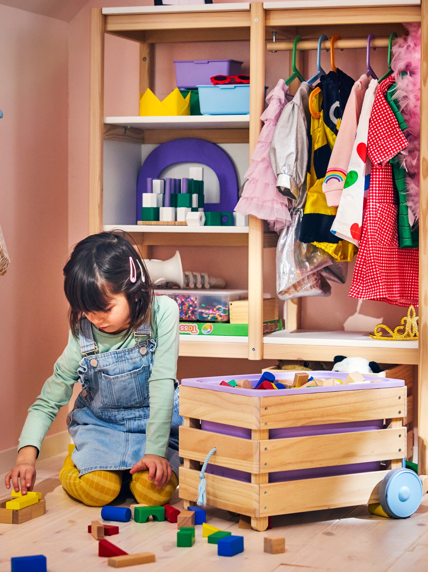 A FLISAT toy storage with wheels filled with toys, beside a wooden wardrobe in a pink children’s room with a child playing.