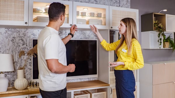 Konsultantka IKEA stojący w żółtej koszuli przy szafce BESTÅ w sklepie IKEA i pokazująca rozwiązania do przechowywania. Przy szafce stoi również klient, który omawia z konsultantką dostępne rozwiązania.