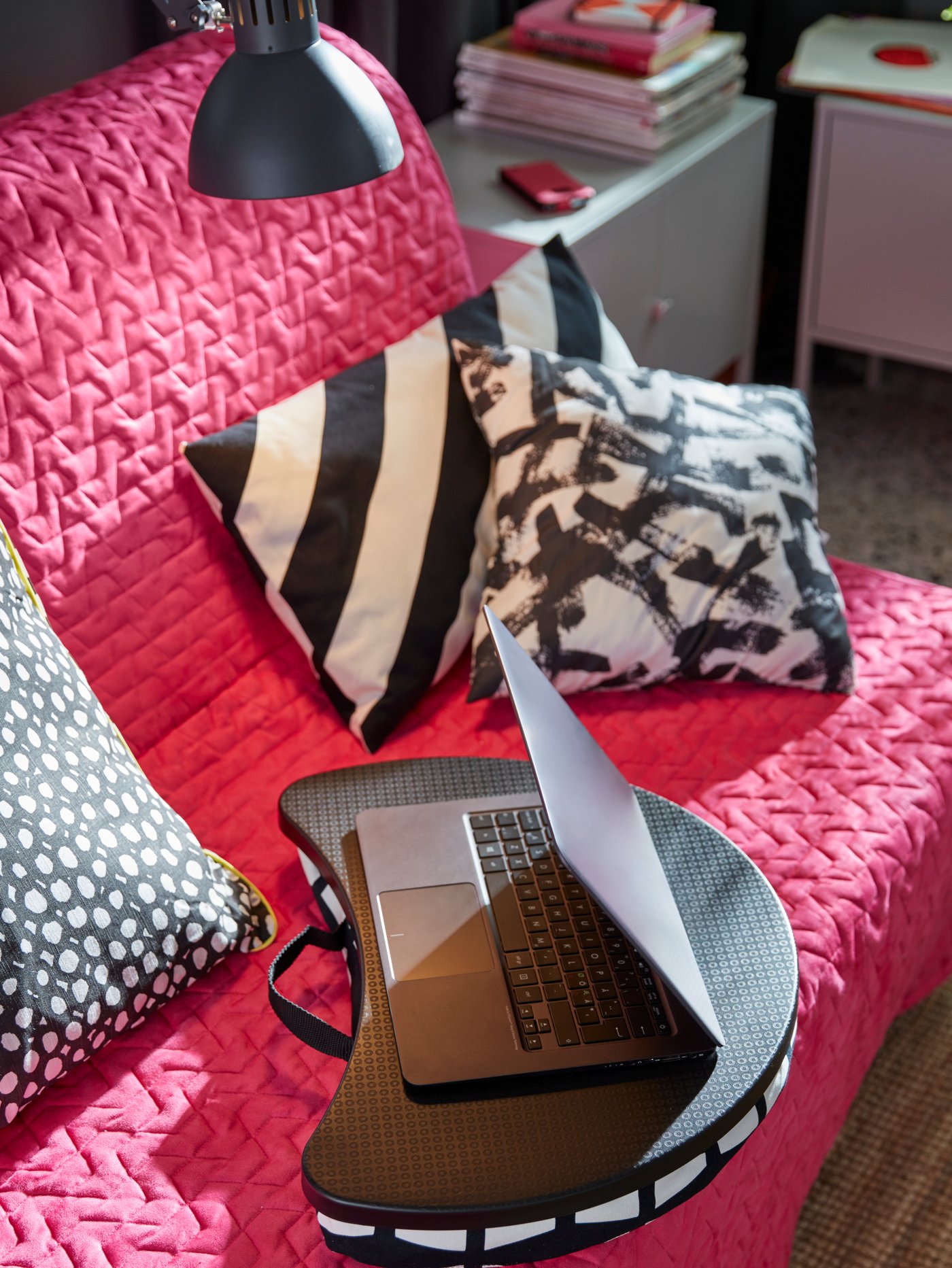 A black and white cushioned IKEA BYLLAN laptop support on a pink sofa amongst grey, black and white cushions.