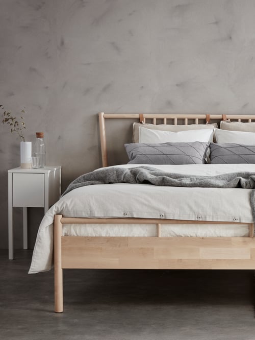 Bedroom with wooden bed frame, duvet cover, pillowcases, bedside table, cushions, carafe with stopper, glass, green plant.