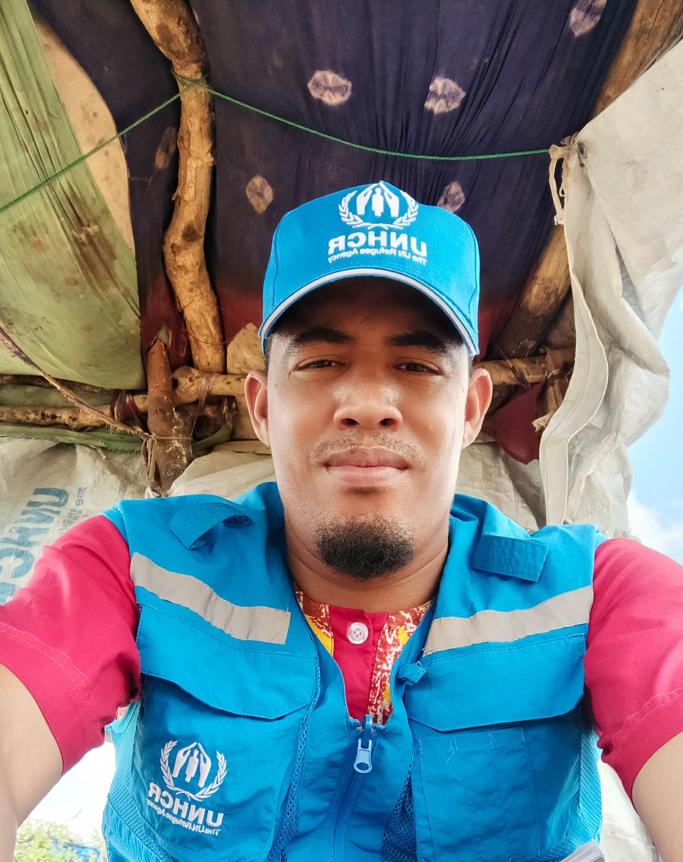 Ilyas outside in a shelter wearing his blue UNHCR work uniform.