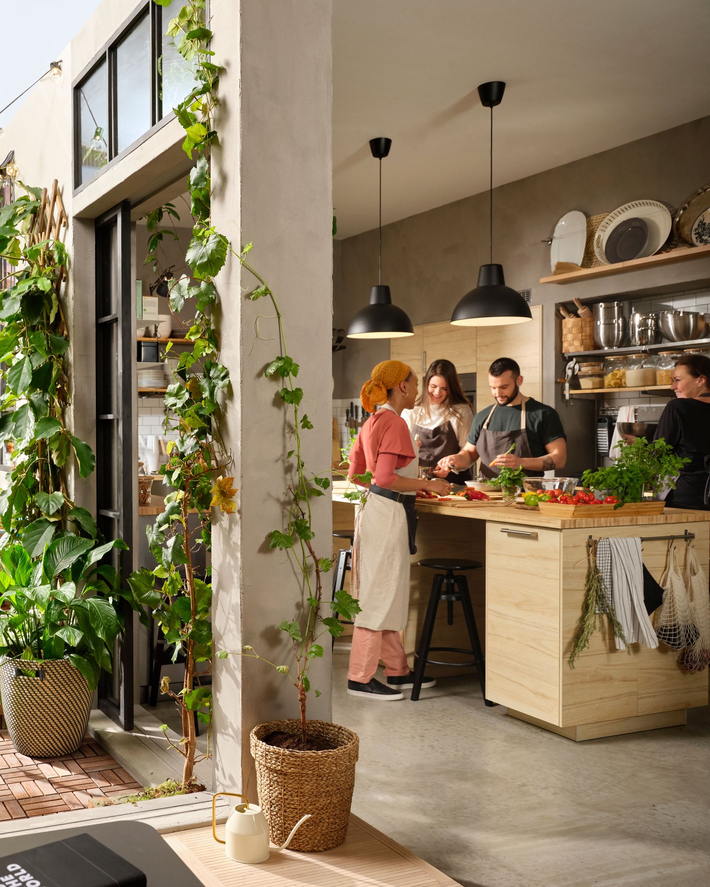 Vier Personen bereiten an einer grossen Kücheninsel Speisen zu. Eine zum Hof geöffnete Tür eröffnet den Blick auf Grünpflanzen.